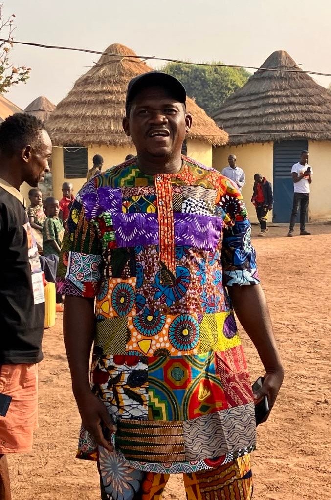 Billy Konaté in farbenfroher Kleidung vor Rundhäusern mit Strohdächern in Guinea