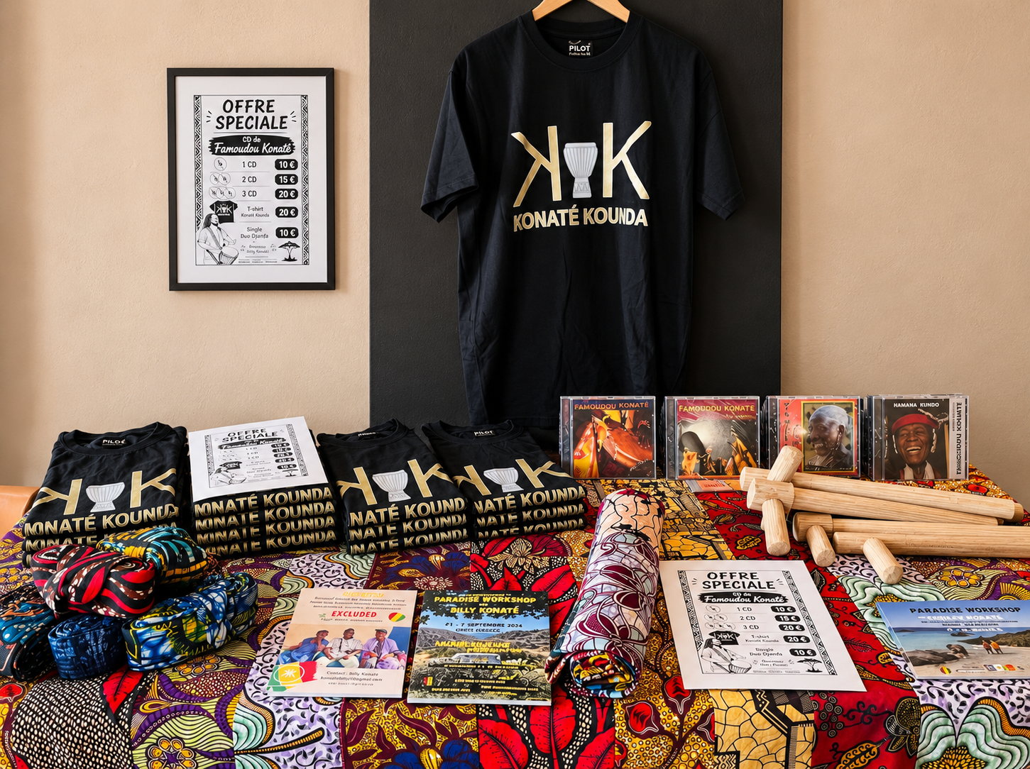 Konaté-Kounda-T-Shirts, CDs, Holzstöcke für Basstrommeln, bunte Gürtel und gemusterte Stoffe auf einem Verkaufstisch