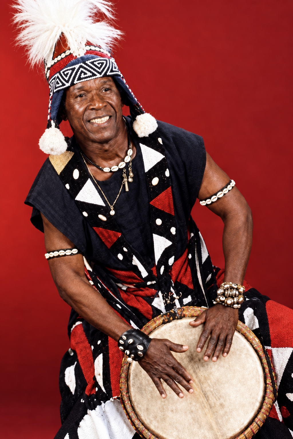 Über mich Famoudou Konaté sitzt in traditioneller Kleidung mit einer Djembe vor rotem Hintergrund