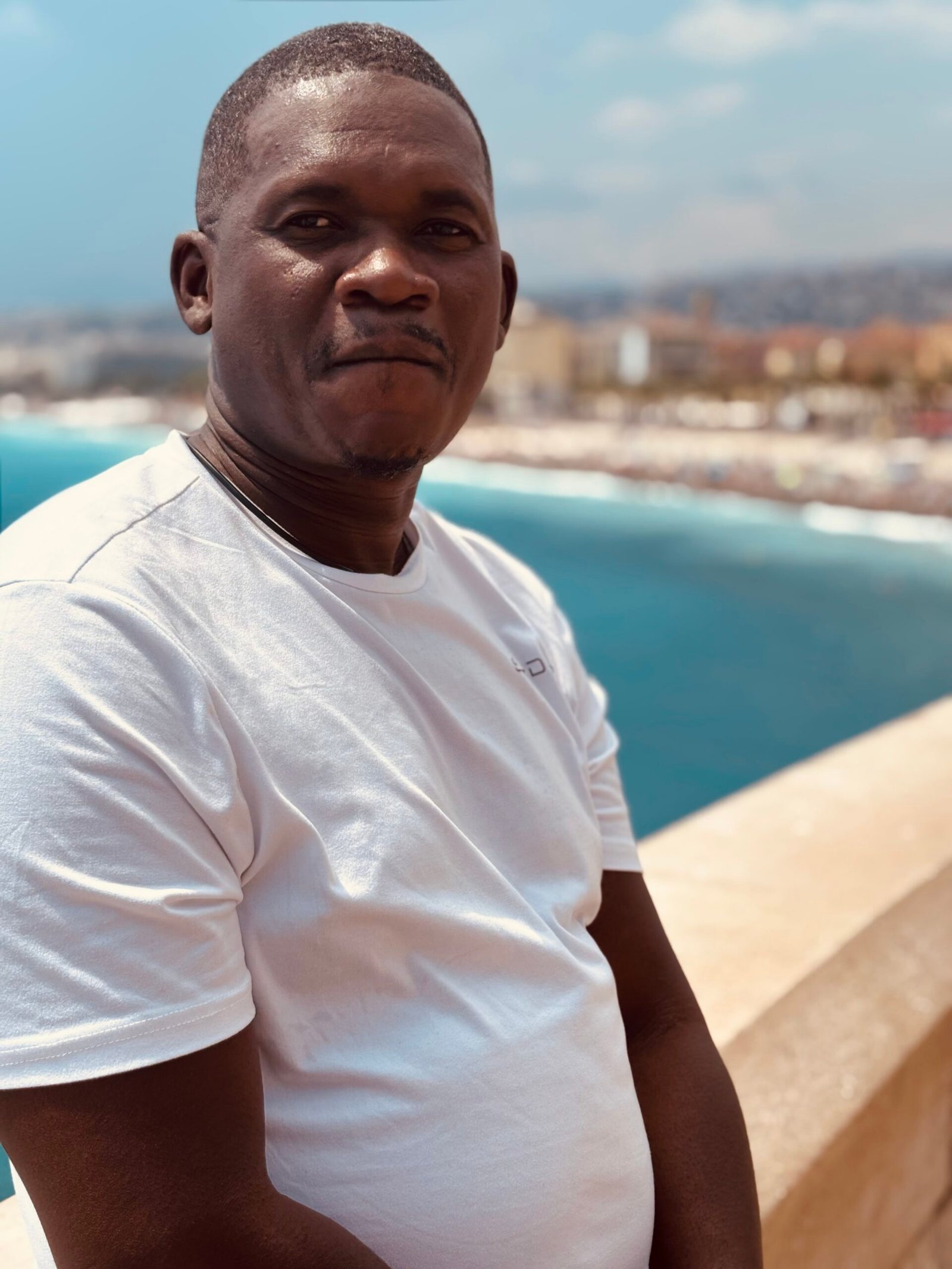 Billy Konaté mit blauer Kappe und Brille vor einer Meeresbucht